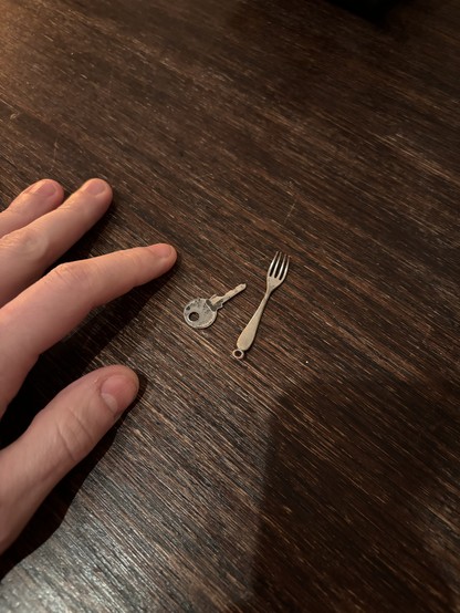 The image features a dark wooden surface with visible grain patterns. A person’s hand, partially visible on the left side, is not the main focus. Positioned on the wooden surface are a small metal key with a traditional design and a miniature fork with four tines and a rounded handle. Soft lighting casts shadows, emphasizing the texture of the wood.
