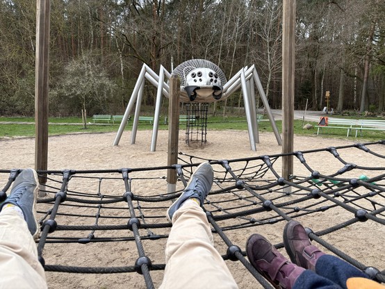 A first-person view of two people’s legs, one wearing beige pants and blue shoes, the other in dark pants and purple shoes, as they lay on a black netted playground equipment. In front of them is a unique playground structure resembling a giant spider with a metal frame and climbing nets, set on a sandy surface surrounded by greenery.