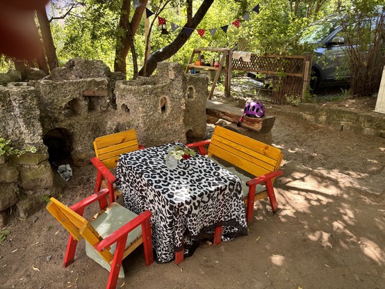 A children’s furniture set with two chairs and a bench arranged around a table with a black/white leopard print tablecloth. Behind the seating area is a self-build castle. In the background is a sand pit and a pretend kitchen.