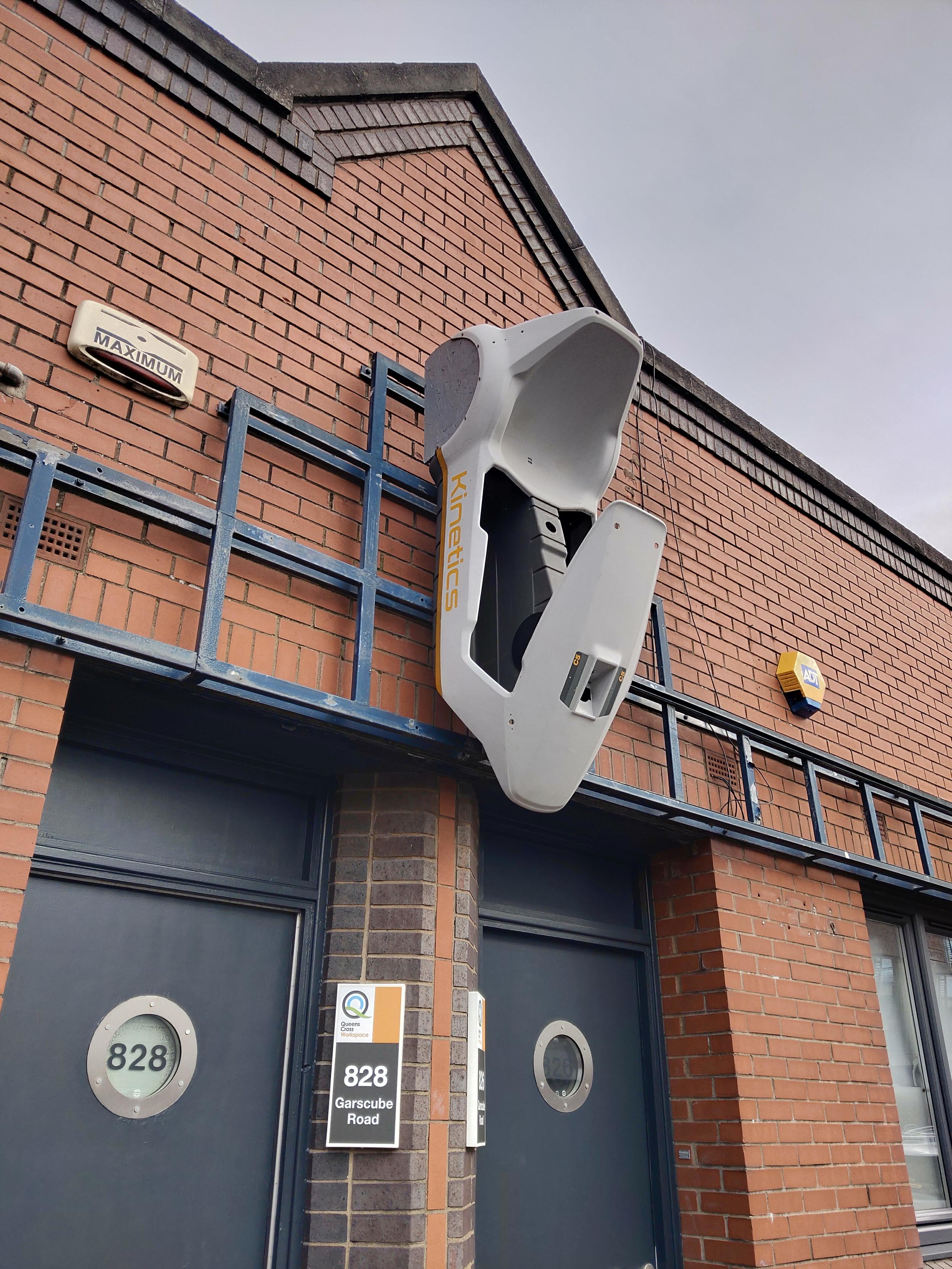 A building with a brick facade and dark blue double doors labeled '828' and '826'. Above the doors, hangs the chassis of a Sinclair C5, a small electric vehicle, mounted on a blue metal frame. The word 'Kinetics' is visible on the chassis. Nearby, a yellow and blue ADT security alarm box is fixed to the wall, and a 'Maximum' security alarm box is visible on the upper left side of the building. A building with a brick facade and dark blue double doors labeled '828' and '826'. Above the doors, hangs the chassis of a Sinclair C5, a small electric vehicle, mounted on a blue metal frame. The word 'Kinetics' is visible on the chassis. Nearby, a yellow and blue ADT security alarm box is fixed to the wall, and a 'Maximum' security alarm box is visible on the upper left side of the building.
