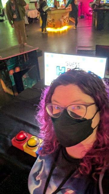 A selfie of me, wearing a mask, standing in front of my game in the Indie Arcade. 