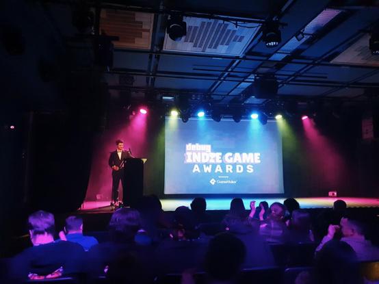 Bright stage, with host behind lecturn and screen showing 'Debug Indie Game Awards'