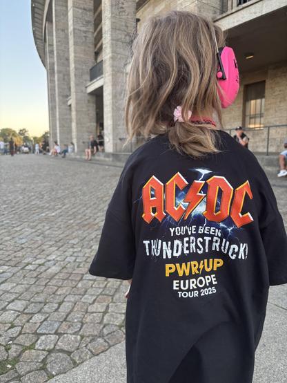 My daughter seen from the back wearing an AC/DC tour shirt with the text AC/DC You’ve been Thunderstruck PWR Up Europe Tour 2025