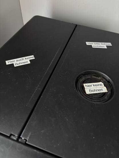 Oberseite der Kaffeemaschine mit zwei geschlossenen Fächern mit dei Aufklebern

“Hier keine Bohnen” und “hier auch keine Bohnen auf Aromalüftung und Wassertank. „hier bitte Bohnen einfüllen” auf dem Bohnenfach