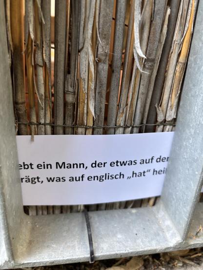 Ein Hinweisschild einer Schnitzeljagd. Es ist am Balkongitter eingeklammt und man kann erkennen „ein Mann, der etwas auf dem“ „trägt, was auf english hat heißt”