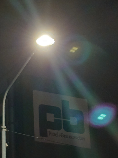 Eerie image of a lamp post in the dark with intense lense flares in front of a building with a large sign saying "pb", which is the logo of a large construction services company.
