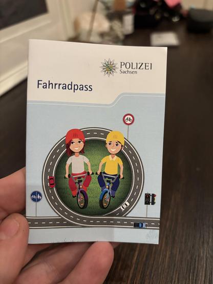 Fahrradpass der Polizei Sachsen Vorderseite. In der Mitte zwei Kinder mit Rad und Helm. Um sie herum verläuft eine Schlaufe aus Straße mit vereinzelten Autos. Es gibt ein Fahrrad-Verboten-Schild, ein Schild für geteilten Fuß- und Radweg sowie eine Ampel