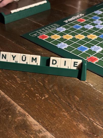 Scrabble board in the background.
In the foreground are nonsensical tiles N Y Ü M and then D I E