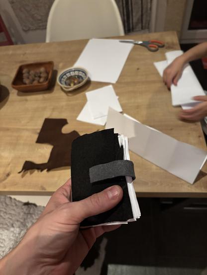 Me holding a felt bound stack of zine-like papers. In the background the kitchen table with more pieces of paper, scissors and my daughter’s hands folding paper.