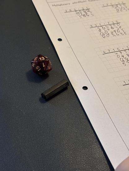 A desk with a sheet of math homework, a cap of a pen and a 20-sided die showing a 13