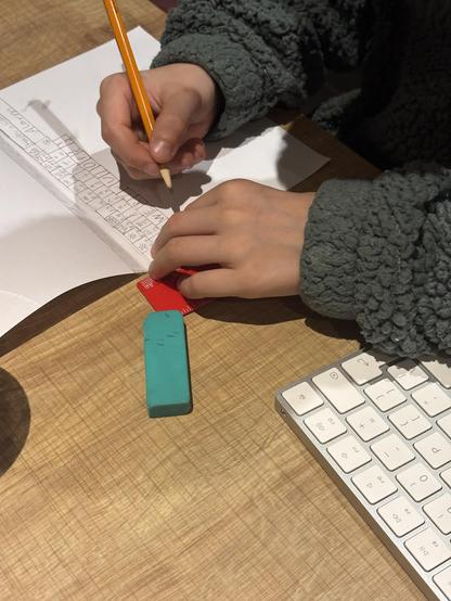 A photo of two kid’s hands with pencil and ruler drawing a keyboard of a folded piece of paper. Next to him is a real Apple keyboard for reference