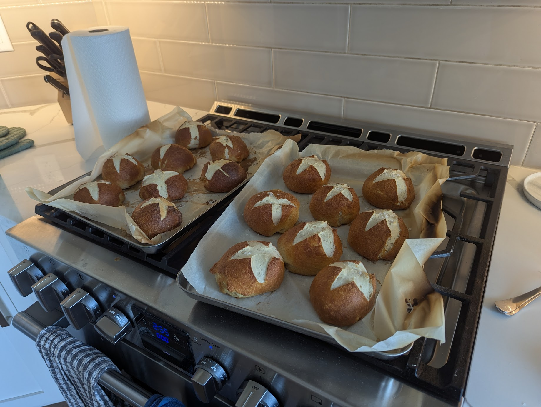 16 golden-brown pretzel buns with a white scored cross on the top of each, sitting on top of a gas stove. You can practically smell the delicious scent of fresh-baked bread through the photo, and feel an overwhelming urge to bake something tasty for your own household.