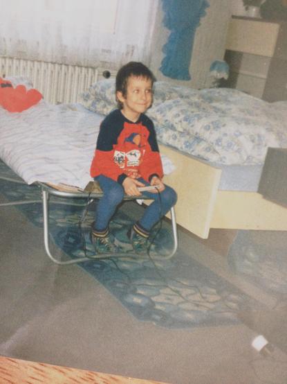 Photo of the post’s author as a child sitting on a guest bed with a NES controller in hand smiling
