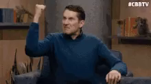 a man in a blue sweater is sitting in a chair and raising his fist .