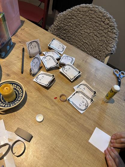 Table from above with printed out gravestones with RIP written on them. Those are then glued to toilet paper rings so they can stand. You can also see glue stick, scissors and my daughters hand putting stuff together