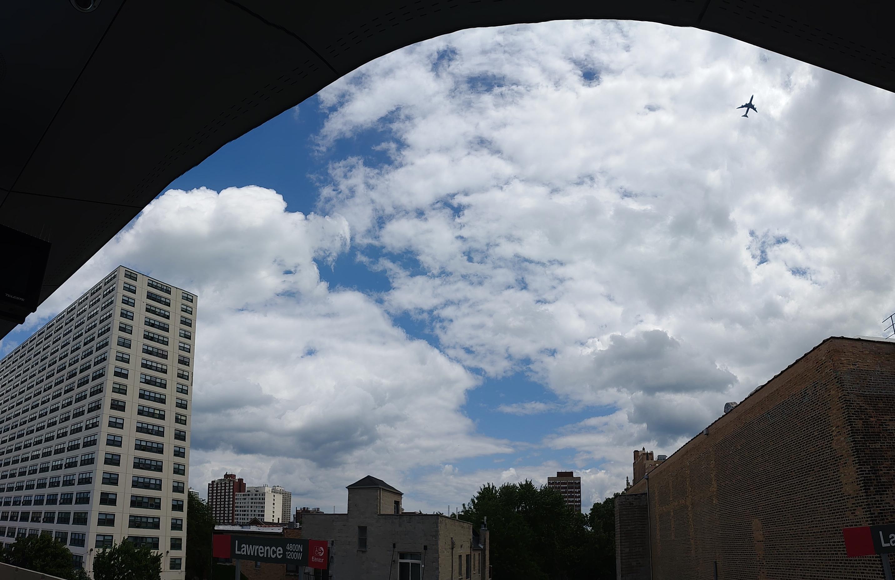 A panorama photo of the sky taken from the lawrence red line stop.  There's an airplane in view, high above.  The clouds are big and fluffy and bright and fill most of the view.