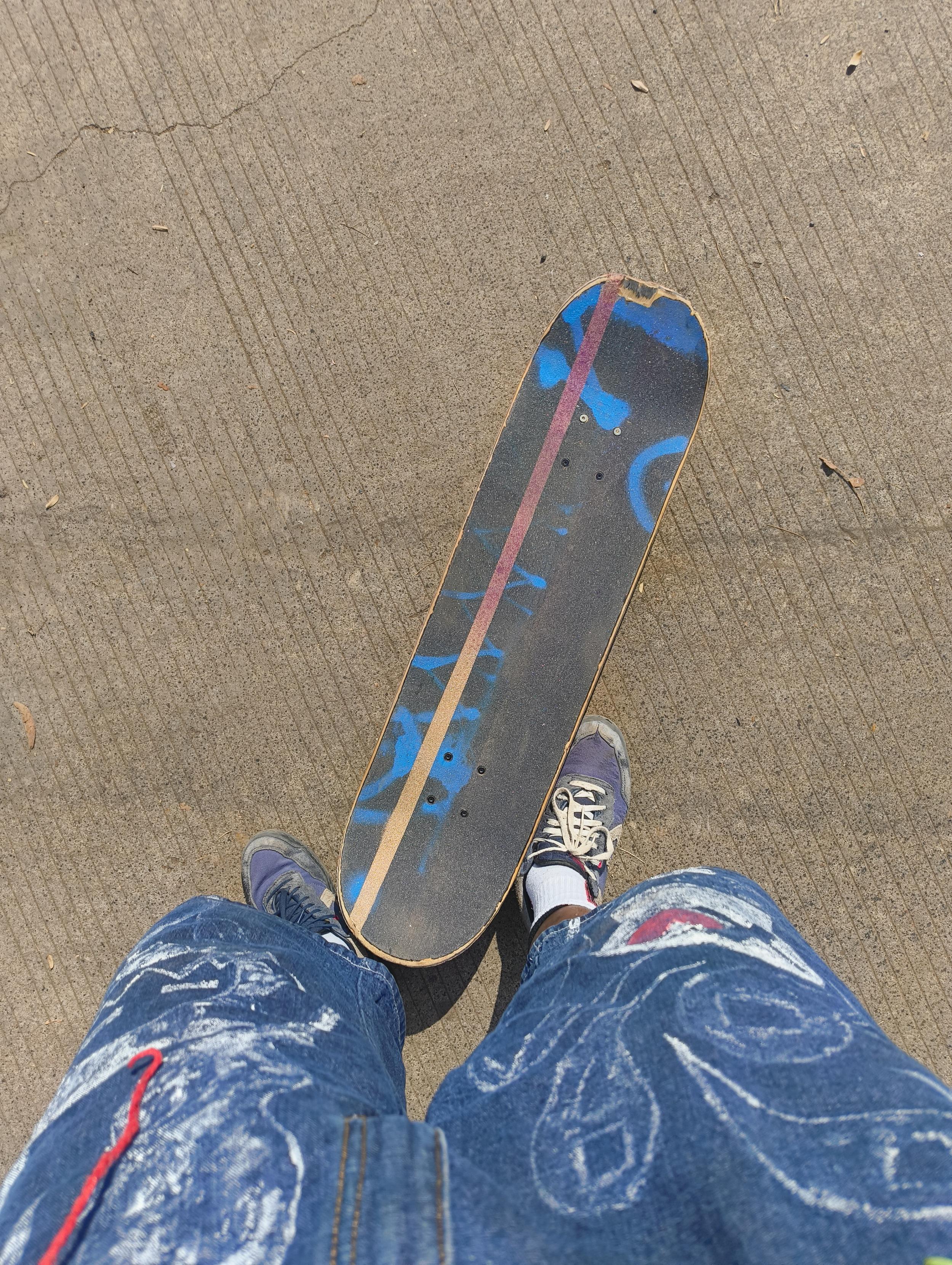 Skateboard deck with a slightly dirty griptape. The griptape has seemingly random blue lines of spraypaint across it, but these mark out spots on my board I pay attention to. There is a single thick gold and purple gradient line that goes across the deck leaning to the left side of it. You can also see my painted denim jorts and worn down converse sneakers