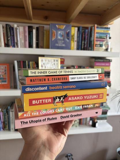 A stack of books from top to bottom:

The Inner Game of Tennis by W. Timothy Gallwey
Shop Class as Soulcraft by Matthew B. Crawford
Discontent by Beatriz Serrano
Butter by Asako Yuzuki
All The Colors Came Out by Kate Fagan
The Utopia Of Rules by David Graeber