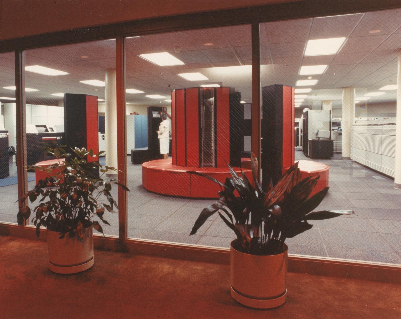Vintage photo of a red CRAY X-MP supercomputer in a computer lab, photographed from the outside with some potted plants and fugly carpeting in the foreground