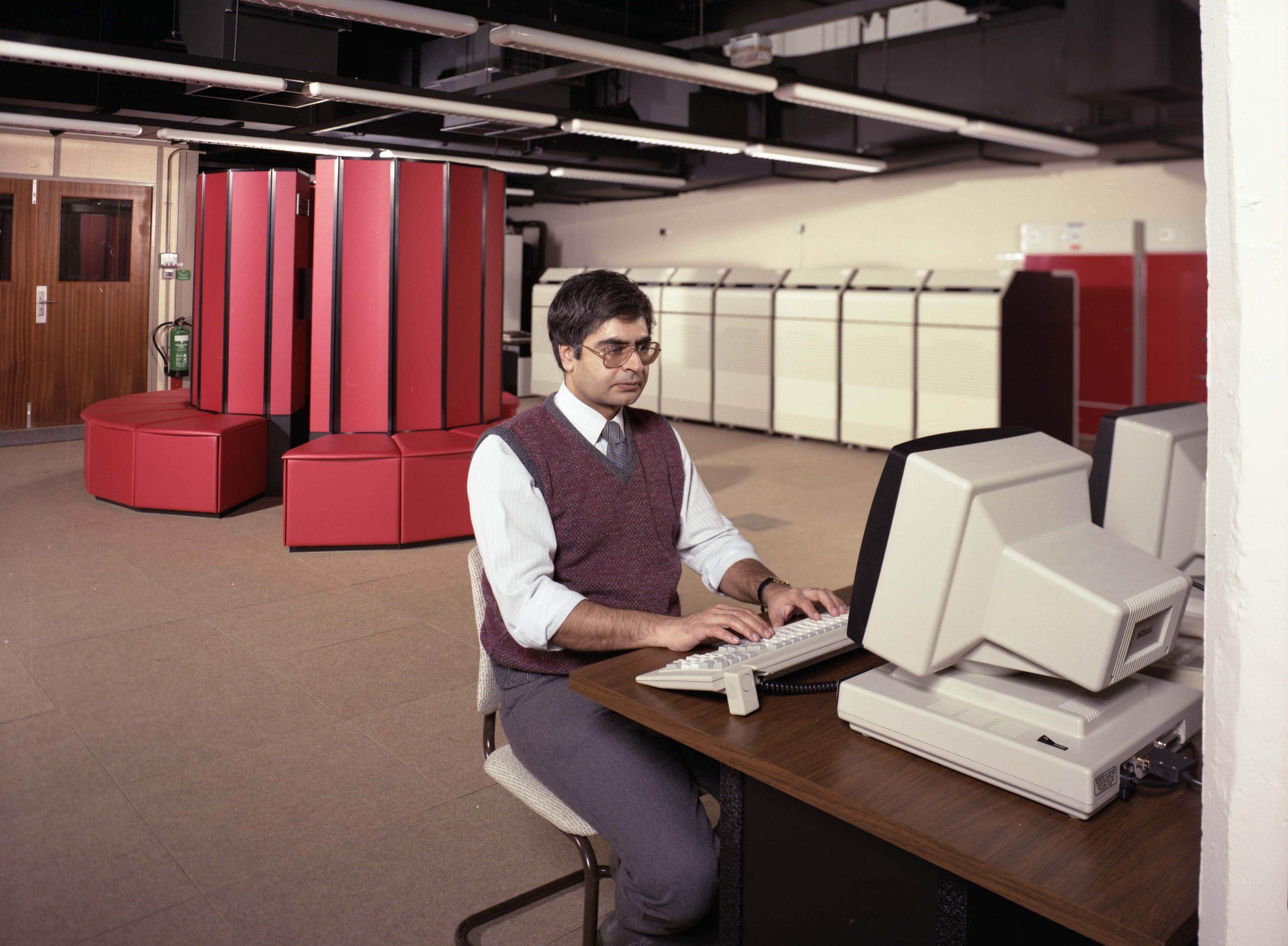 Photo of a person sitting in a computer lab at a keyboard and computer screen, typing. IN the background is a red CRAY X-MP supercomputer