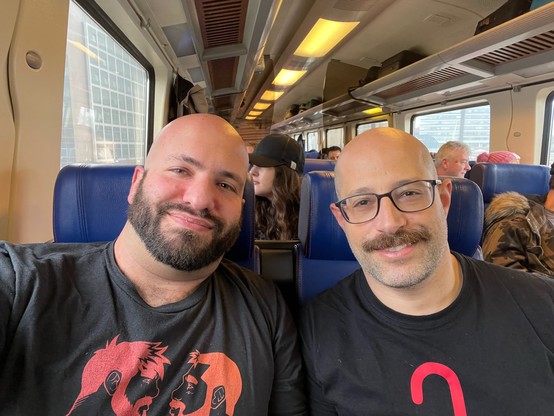 Two men sitting side by side in a train. Both are bald with facial hair, wearing black shirts. I’m on the left, @BoyBrave@woof.group on the right.