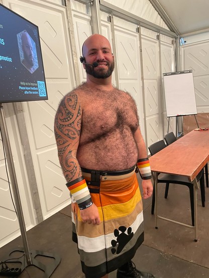 Me preparing for the Masterclass. I’m wearing my bear flag leather kilt, with bear flag gauntlets, and I’m shirtless, showing off my hairy chest and my Polynesian tattoo on my right arm. I’m wearing a head-mounted microphone, and I’m looking at the camera and smiling. Behind me is a table, flipchart, screen, and white walls.