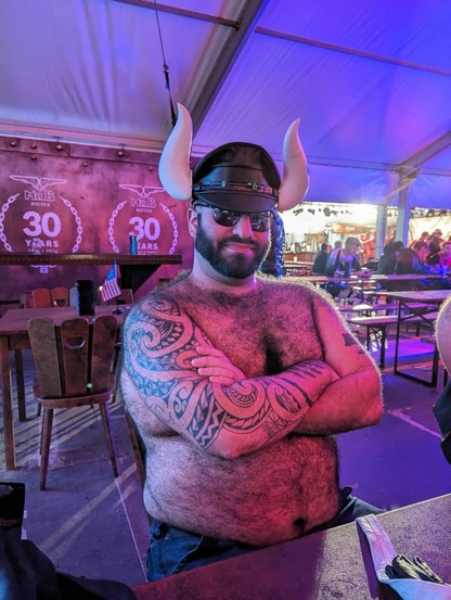 Me wearing sunglasses and a Muir cap with large white bull horns affixed to the sides. I’m shirtless, with my arms folded in front of my hairy chest, with a smirk on my face.
