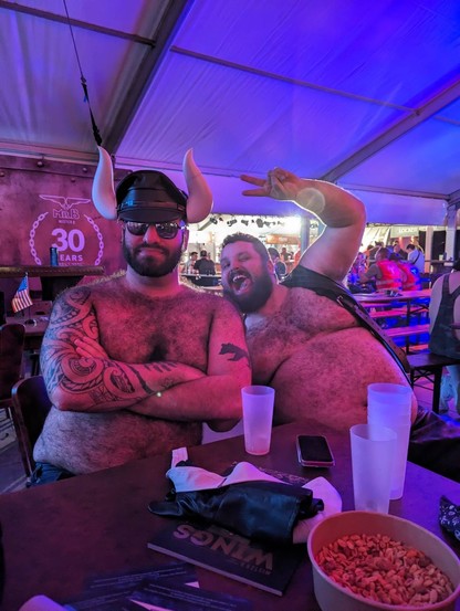 The same picture, with @lthrbearsolid leaning in with a giant cheesy smile and fingers in a two-finger “peace” pose. He is also shirtless, wearing a leather bar vest.