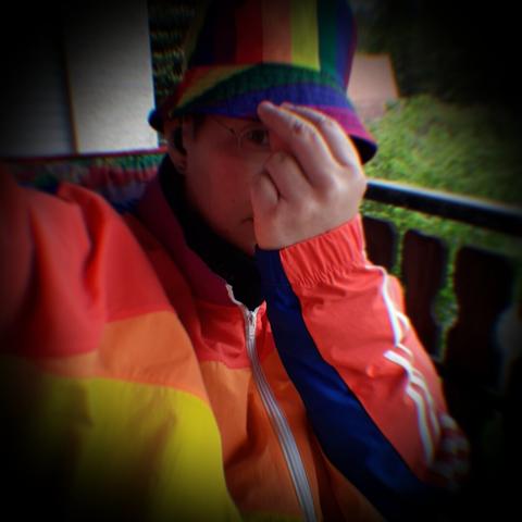 Eine weiße Person mit Regenbogenhut und regenbogenbunter Jacke sitzt auf dem Balkon. Das Gesicht ist von der linken Hand verdeckt, die an die Hutkrempe greift.