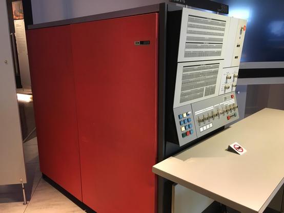 Photograph of the front panel of an IBM System/360 mainframe—an adult person height red box about 1.5 metres deep, with a panel of blinkenlights and rotary dials (and some buttons). A desk sits in front of it, presumably for the operator's teletype printer.