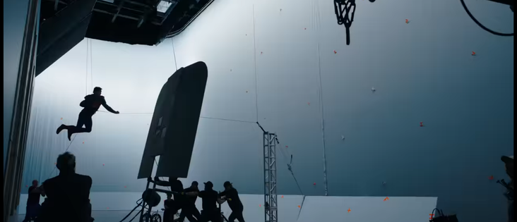 Screen cap from "Superman Day" behind the scenes YouTube video, where is appears the silhouette of Superman, sans full cape, is rigged with wires and is flying in front of a greenscreen, but the green has been removed for the PR video.  The sillhouette of James Gunn, watching the performance, in in the foreground.