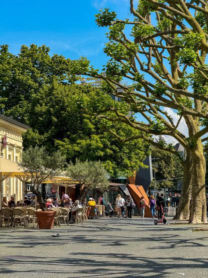 People relaxing at an outdoor café in a sunny city square with trees and modern art sculptures.