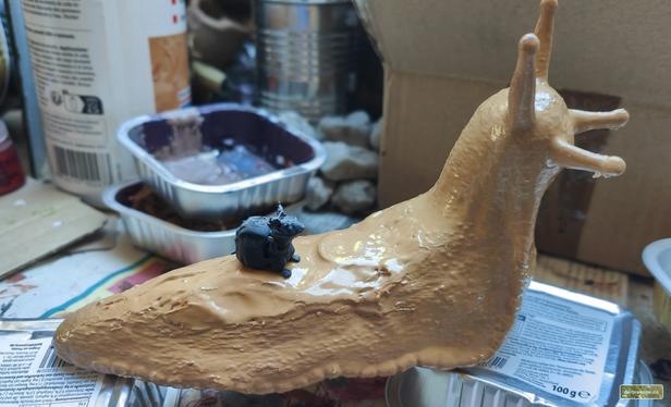 Eine 3D-gedruckte Riesenschnecke  mit einer kleinen Mäuseminiatur auf dem Rücken. Im unscharfen Hintergrund diverse Bastelutensilien.
