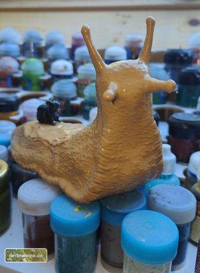 Frontalansicht einer 3D-gedruckten Riesenschnecke  mit einer kleinen Mäuseminiatur auf dem Rücken. Im unscharfen Hintergrund viele kleine Farbnäpfe.