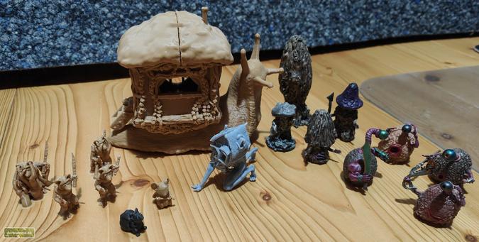 Ein Dutzend gebastelte und 3d-gedruckte Rollenspielminiaturen steht auf dem Holzboden: In der Mitte eine übergroße Schnecke mit einem Holzhäuschen auf dem Rücken, links einige Ratten und Mäuse. Rechts hinten vier Pilzartige Wesen und rechts vorn vier Wesen, die den Buchlingen aus Walter Moers Zamonienromanen nachempfunden sind.
