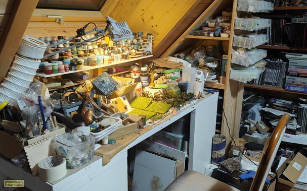 Ein überquellender Basteltisch mit ungezählten Projekten in verschiedensten Stadien der Fertigstellung. Im Hintergrund ein Bücherregal voller Rollenspielbücher.