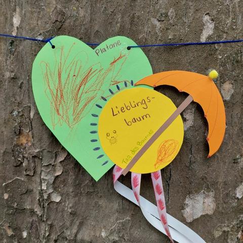 Eine umfangreiche Platane hat einen Orden um den Stamm gewickelt: ein grünes Herz, auf dem seitlich ein gelber Kreis klebt mit der Inschrift "Lieblingsbaum, Tag des Baumes". Ein oranger Sonnenschirm ist auf den Kreis gebastelt. Am Orden hängen Flatterbänder.