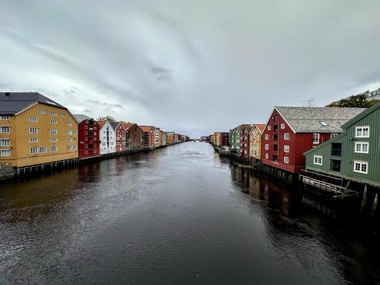 This image shows a row of colorful wooden buildings directly along the edge of a river. These buildings are known as 'stilt houses,' standing on pilings over the water. The architectural style and the arrangement suggest they are likely located in a Northern European country, possibly Norway. The overcast sky and the calm water create a tranquil and slightly moody atmosphere. The buildings are painted in various shades, including red, yellow, and green, which are typical for historical buildings in some Scandinavian waterfronts. This scene might be found in a city like Trondheim, where such waterfront architecture is iconic.