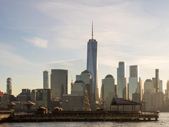 Golden hour view of Lower Manhattan skyline from Paulus Hook waterfront in Jersey City, featuring One World Trade Center and a decorated Christmas tree on the pier.