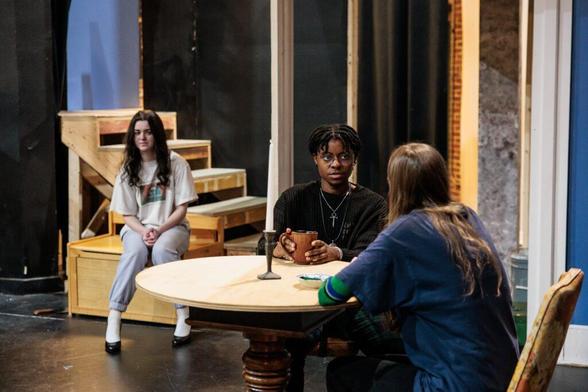 Three people on a stage set up like a house interior. There are two women and a man. One woman sits on a chest by the stairs, while the man and other woman are witting at a round table, drinking out of mugs and talking.