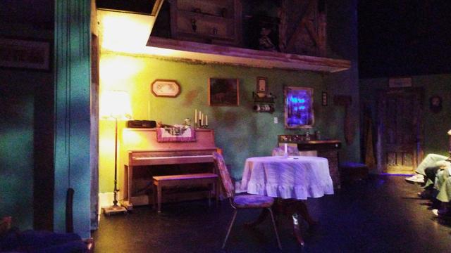 A view of the set on a stage. There is a table and chairs, a piano, an upstairs area, and a window with strange blue lighting outside. The interior is colorful yet dark, and has a ominous feel.