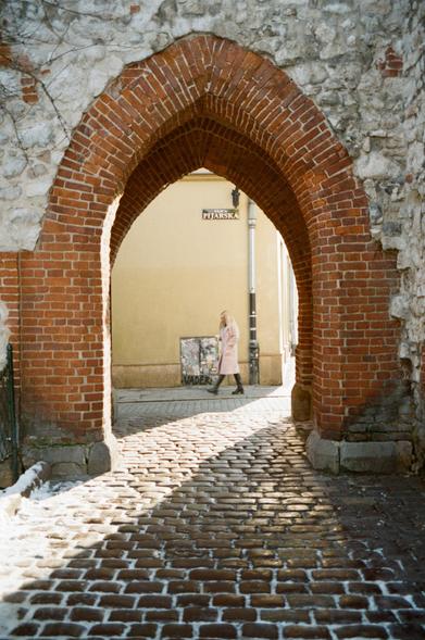 Colour photo of an archway in old town Krakow, poland. Taken in January 2025, there is still some snow on the cobble stones. The archway is red brick within a white stone wall. A lady in a white coat passes in view of the arch as I'm taking the photo.