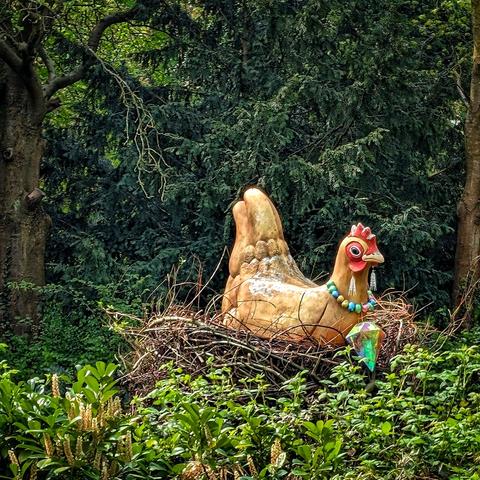 The picture shows a whimsical scene featuring a large, light-brown ceramic chicken sitting in a nest made of twigs. The chicken is adorned with a colorful beaded necklace of blue, turquoise, yellow, and orange. It has a bright red comb and wattle, and a curious expression with its black button eye looking to the right.
Adding to the fantastical nature of the scene, a large, multi-faceted gemstone, predominantly green and clear, is positioned in front of the nest, seemingly placed there. The nest itself is nestled amongst lush green foliage and small white flowers, suggesting an outdoor setting, perhaps a garden or a whimsical park. Behind the nest and chicken, a backdrop of dark green trees and glimpses of branches create a natural, slightly shadowy environment, making the brightly colored chicken and gemstone stand out. The overall impression is one of playful artistry and unexpected charm.