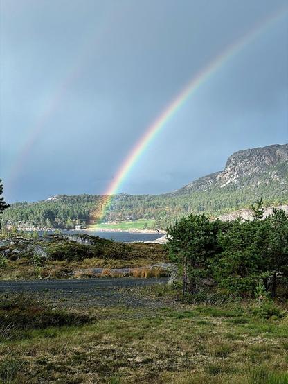 This image captures a beautiful natural landscape featuring a double rainbow arcing through a cloudy sky. The foreground includes a grassy field with scattered trees and shrubs, while the middle ground reveals a body of water partially enclosed by land. Beyond the water, there are prominent rocky hills or low mountains, which contribute to the scenic beauty of the area. The rainbows appear to touch down on the far side of the water, adding a striking splash of color to the scene. Overall, the composition conveys a serene and picturesque setting, likely after a rain shower given the presence of rainbows and wet surfaces.
