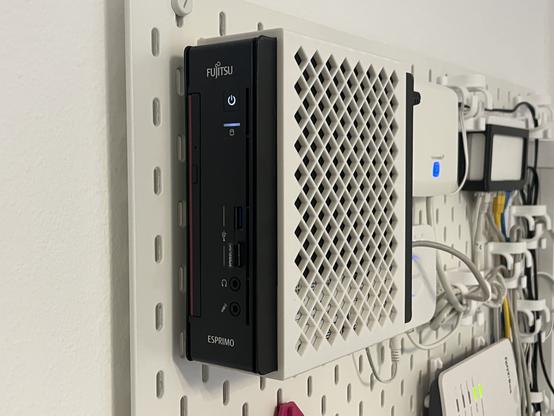 Das Bild zeigt ein schwarzes Fujitsu-ESPRIMO-Computer-System, das an einer weißen Lochplattemontiert ist. Das Gerät hat eine schlanke, vertikale Form und ist mit verschiedenen Anschlüssen ausgestattet, darunter USB-Ports und Audioausgänge. Es ist mit einem Netzteil verbunden, das durch ein weißes Gittergehäuse mit einem Lattice-Muster abgedeckt ist.. Die Lochplatte ist mit verschiedenen Kabeln und Zubehörteilen wie Kabelhaltern und anderen Geräten wie einem Router versehen. Die Umgebung wirkt ordentlich und organisiert, was auf eine professionelle oder technische Umgebung hinweist.