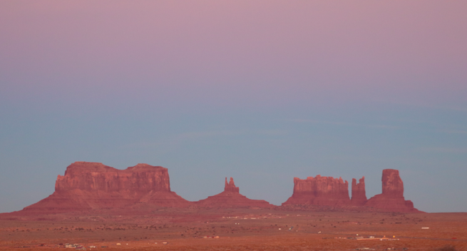 massive sandstone buttes can be seen rising from the desert floor under a dusky, cloudless, pastel sky as the sun sets