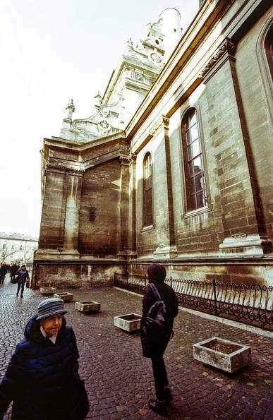This is a film photograph taken in Lviv, Ukraine. The image shows a group of people walking along a cobblestone street next to a grand historical building, likely a church or cathedral. The structure features tall arched windows, ornate stonework, and classical architectural details, such as columns and sculptures adorning the upper facade. The lighting gives a slightly faded, vintage feel characteristic of analog photography. The atmosphere is calm, and the people are dressed warmly, suggesting it might be autumn or winter.