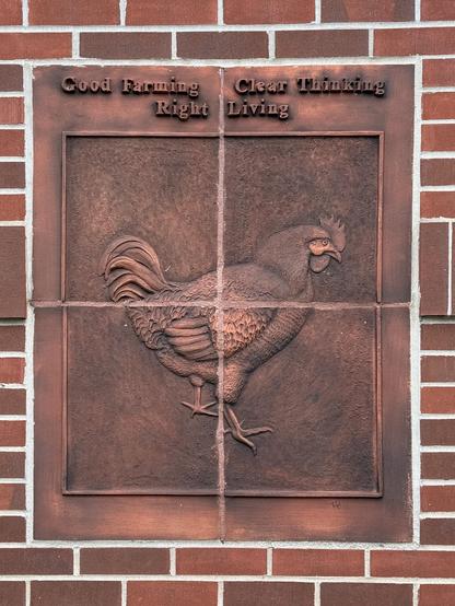 Four square clay tiles arranged to form a larger square surrounded by bricks in a wall. The tiles each contain a quadrant of a relief of a chicken. Relief text above the chicken reads “Good Farming, Clear Thinking, Right Living”