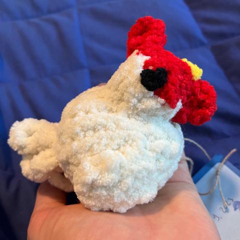 A small chicken knitted out of fuzzy chenille yarn. She has big black embroidered eyes, a red comb and wattle, and a yellow little beak. She is white with a fluffy tail and wings. She has a nametag attached to her.