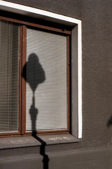 blick auf ein doppelfenster im erdgeschoss in der prallen sonne, die rollos sind geschlossen und der harte schatten einer straßenlaterne legt sich auf das fenster